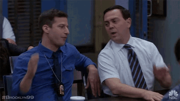 two men are sitting at a table with the hashtag #brooklyn99 on the bottom right