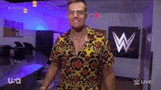 a man wearing sunglasses and a floral shirt is standing in a room with a sign that says wwe live .