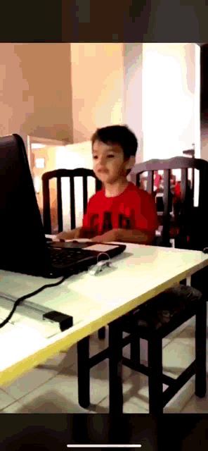 a young boy in a red gap shirt sits in front of a laptop computer