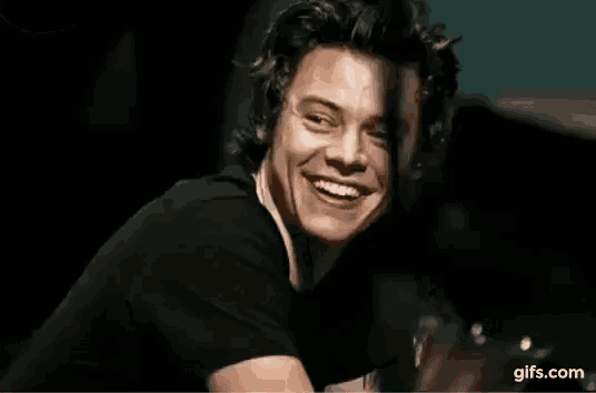 harry styles is smiling and looking at the camera while wearing a black t-shirt .