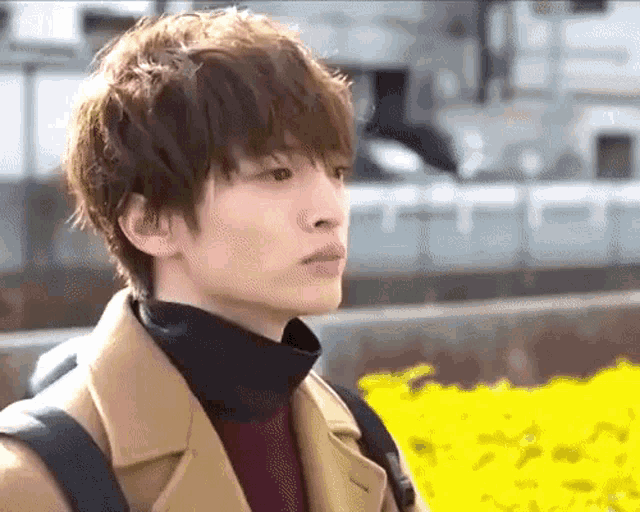a young man wearing a brown coat and a black turtleneck is standing in front of a yellow flower bed .