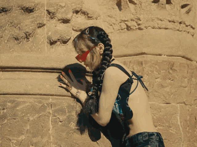 a woman in a braided top and jeans stands in front of a stone wall