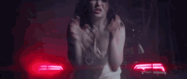 a woman in a white dress is standing in front of a red car with the lights on .