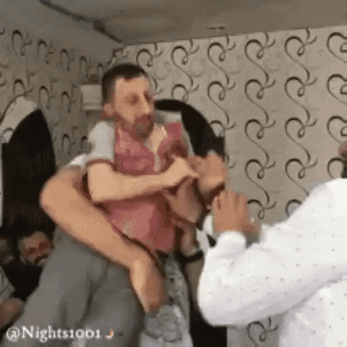 a man is holding a baby in his arms in a room while another man looks on .
