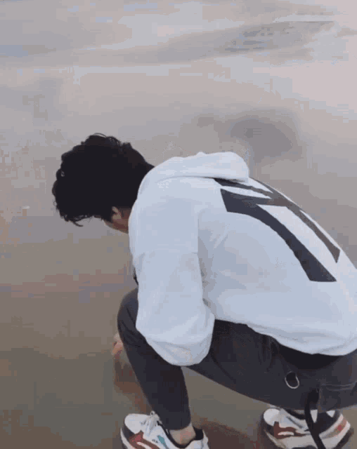a person wearing a white hoodie with the number 7 on the back squatting on the beach