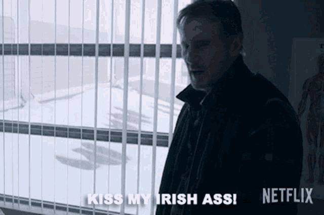 a man in a black jacket is standing in front of a window with blinds and says kiss my irish ass .