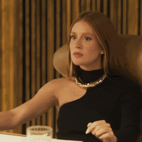 a woman wearing a black one shoulder top and a gold necklace is sitting in a chair .