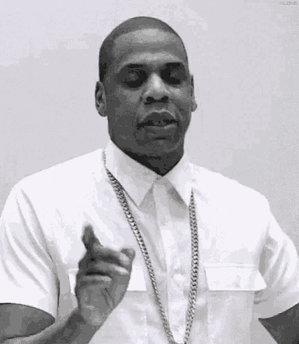 a man wearing a white shirt and a gold chain is pointing at the camera .