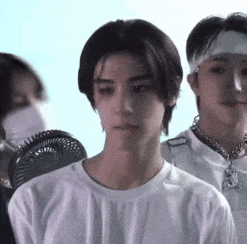a man in a white shirt with a bandana on his head stands in front of a fan