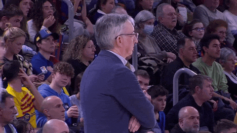 a man with glasses stands in a crowd of people watching a basketball game
