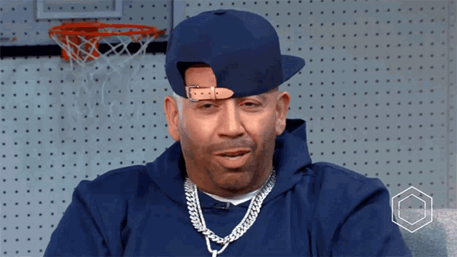 a man wearing a blue hat and a chain around his neck is sitting in front of a basketball hoop