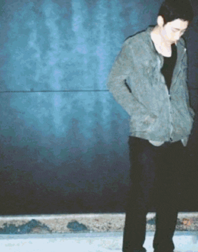 a man wearing a grey jacket and black pants stands in front of a blue wall