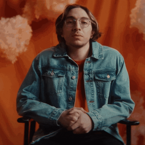 a man wearing a denim jacket and glasses sits with his hands folded