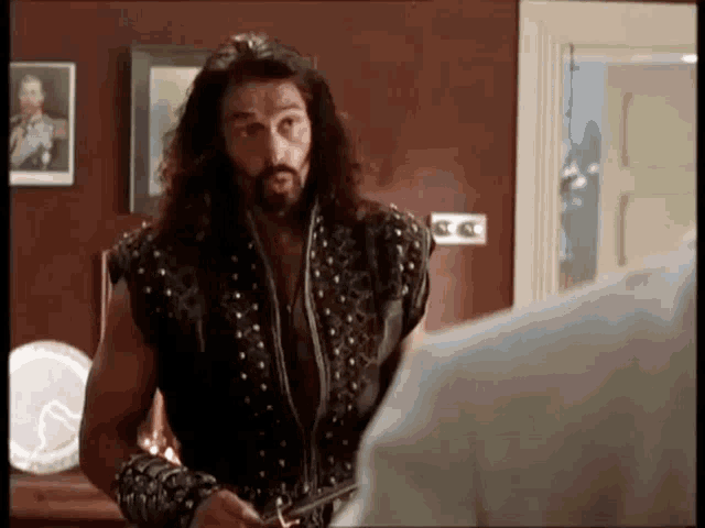 a man with long hair and a beard is holding a sword and talking to another man .