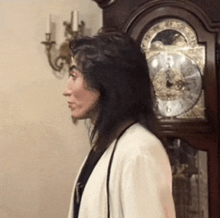 a woman in a white jacket stands in front of a grandfather clock with the hands on the numbers 1 and 2