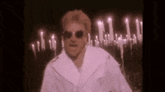 a man in a white coat and sunglasses is standing in front of a row of lit candles .