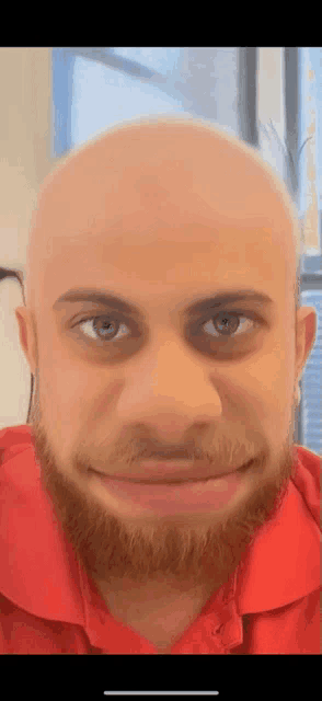 a bald man with a beard wearing a red shirt makes a funny face