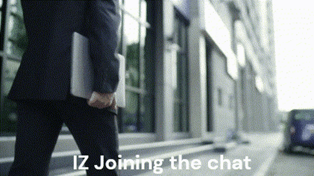 a man in a suit is walking down a street carrying a laptop and the words iz joining the chat below him