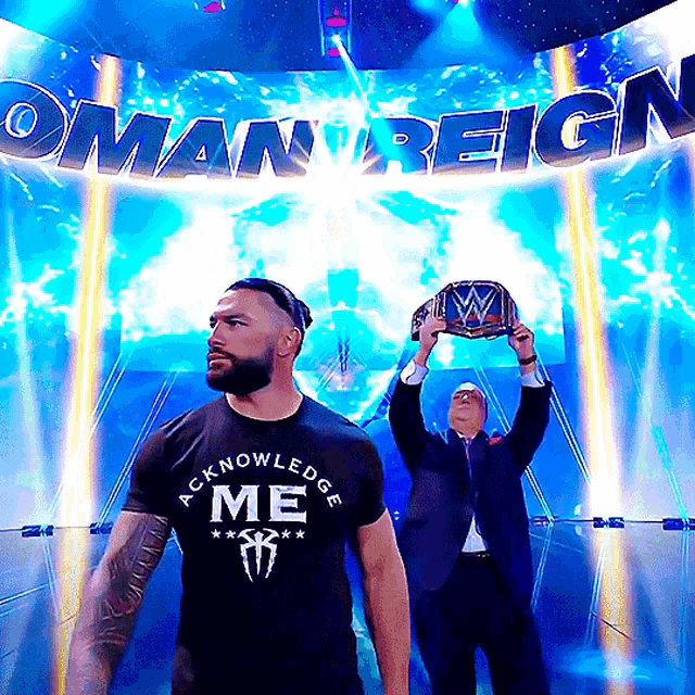 a man wearing a black shirt that says acknowledge me stands in front of a sign that says oman reign