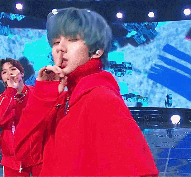 a man in a red hoodie holds his finger to his mouth while singing into a microphone