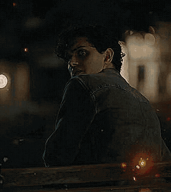 a man in a denim jacket is looking over a wooden railing at night .
