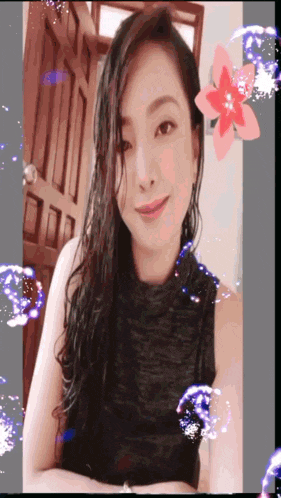 a woman in a black tank top is smiling in front of a door with a flower in the background
