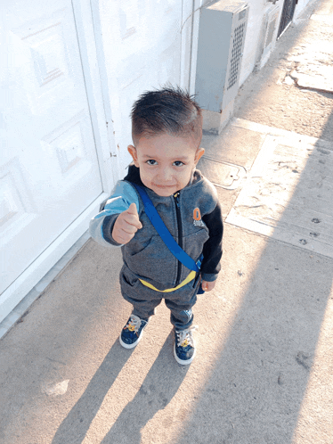 a young boy wearing a gray jacket with a blue strap around his waist is giving a thumbs up