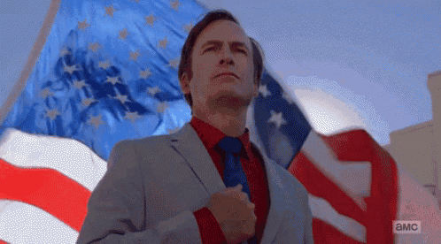 a man in a suit and tie is standing in front of an american flag with his hand on his chest .