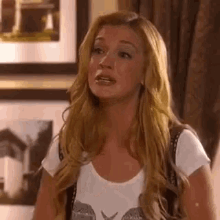a woman with long blonde hair is sitting in a living room with a picture on the wall behind her .