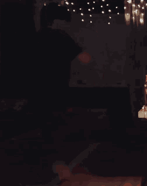 a man in a black shirt and plaid pants is dancing in a dark room with christmas lights hanging from the ceiling .