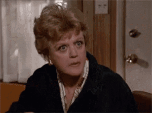 a woman is sitting at a table in front of a door making a surprised face .