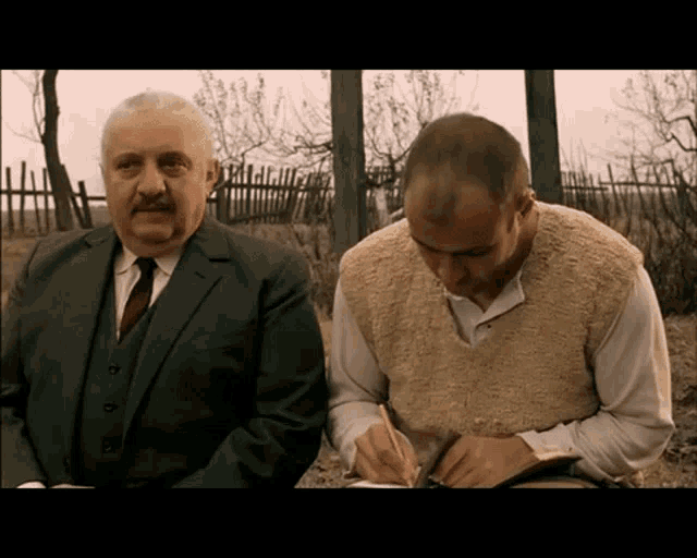 a man in a suit and tie sits next to another man who is writing on a piece of paper