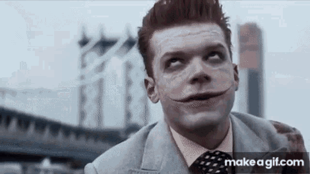 a man in a suit and tie with a joker mask on his face is standing in front of a bridge .