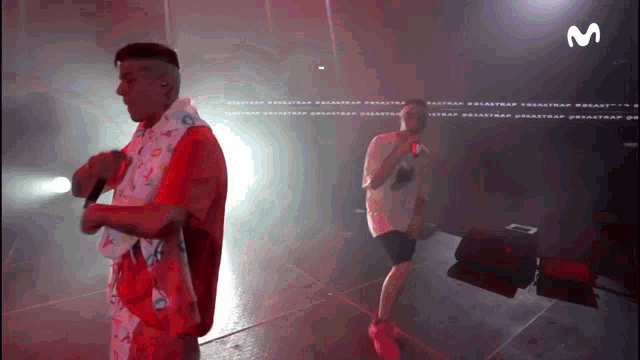 a man in a red shirt is singing into a microphone while another man stands behind him