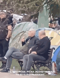 two men are sitting on a bench in front of tents and smoking cigarettes .