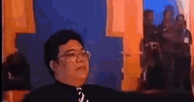 a man wearing glasses and a black shirt and tie is sitting in front of a blue wall .
