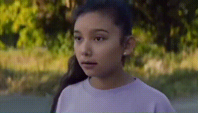 a young girl in a purple shirt is standing in a park looking at the camera .