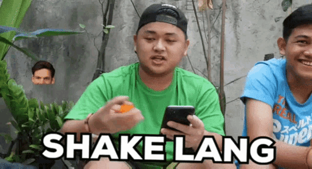a man in a green shirt is holding a cell phone while another man in a blue shirt looks on ..