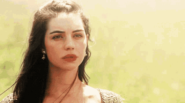 a woman with long dark hair and earrings looks at the camera with a serious look on her face