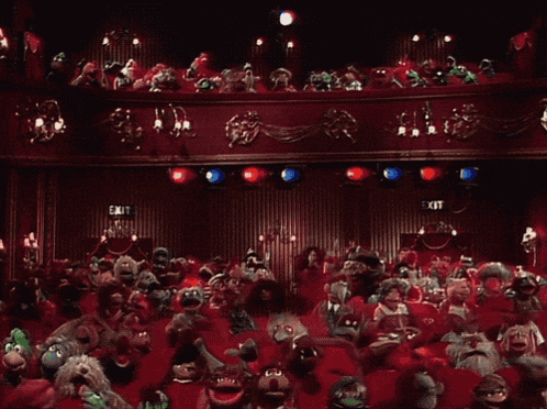 a large group of muppets are sitting in a theater with an exit sign above them