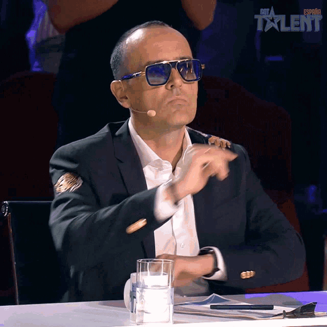 a man wearing sunglasses and a suit sits at a table with a glass of water and a sign that says get a talent