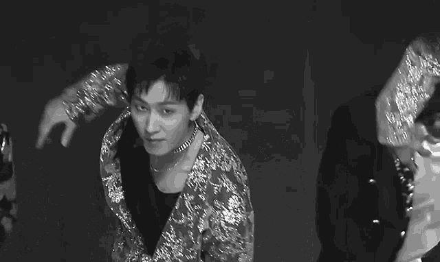 a black and white photo of a man wearing a sequined jacket and a necklace .