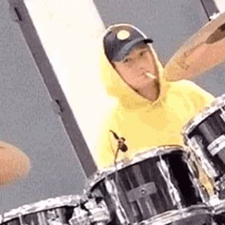 a man in a yellow hoodie is smoking a cigarette while playing drums .