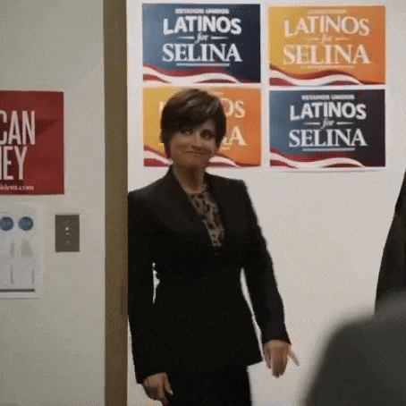 a woman in a black suit stands in front of a wall of posters for latinos for selina