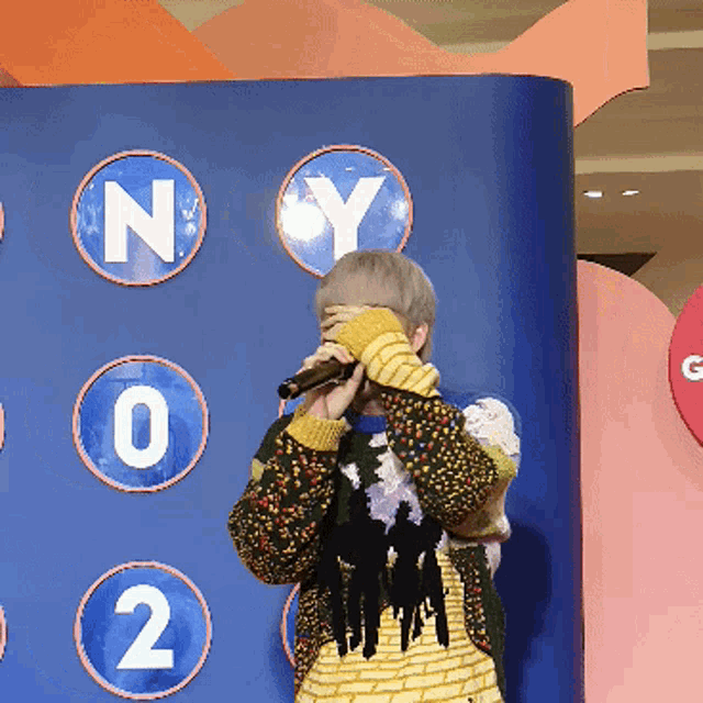 a person covering their face with a microphone in front of a blue wall with ny circles