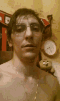 a shirtless man wearing glasses looks at the camera with a clock in the background