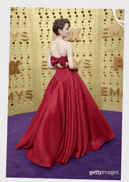 a woman in a red dress stands on a purple carpet in front of a wall that says emmys