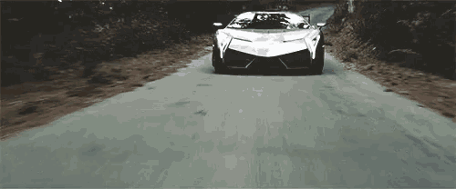 a white sports car is driving down a road in the woods .