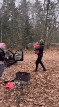 a woman is holding a baby in her arms while standing next to a car in a forest .