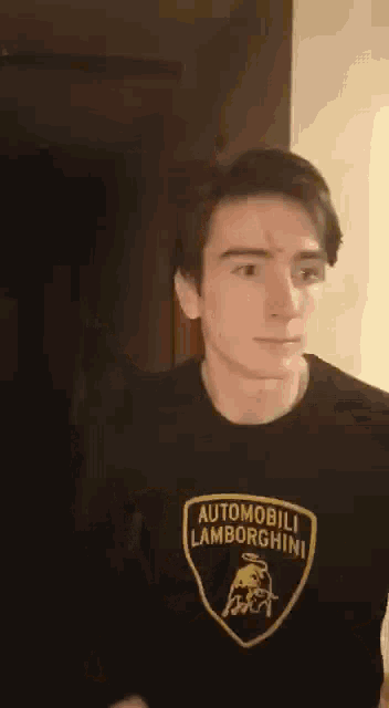 a young man wearing a black t-shirt with a lamborghini logo on it is standing in a dark room .
